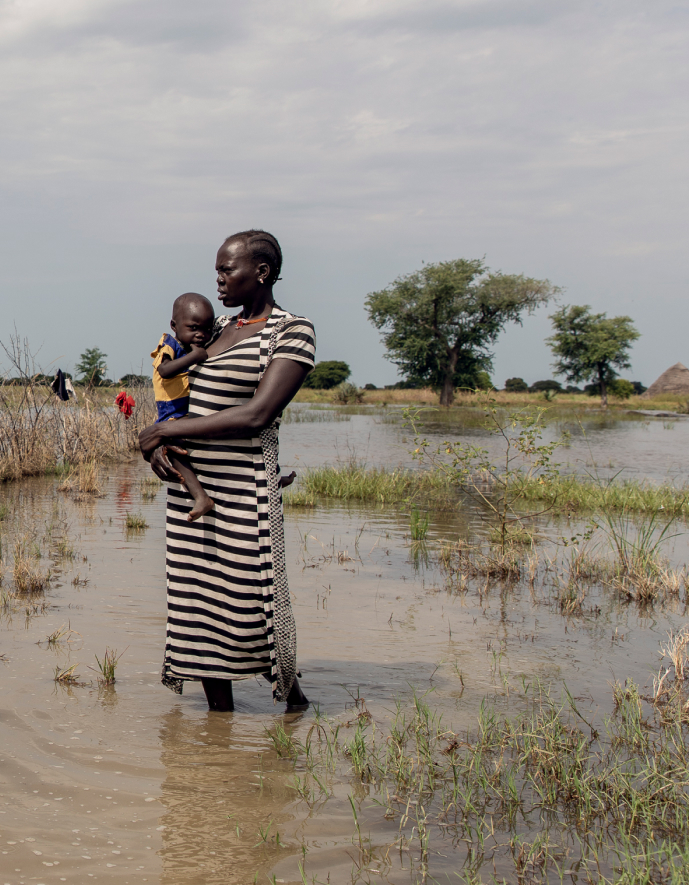 Abuk, holding a sleepy Nyirou, in front of their flooded house, in Northern Bahr El Ghazal, South Sudan.